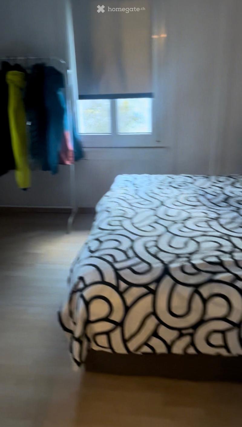 Bedroom with a bed covered in a black and white patterned duvet, a wardrobe with hanging clothes, and a window with blinds.