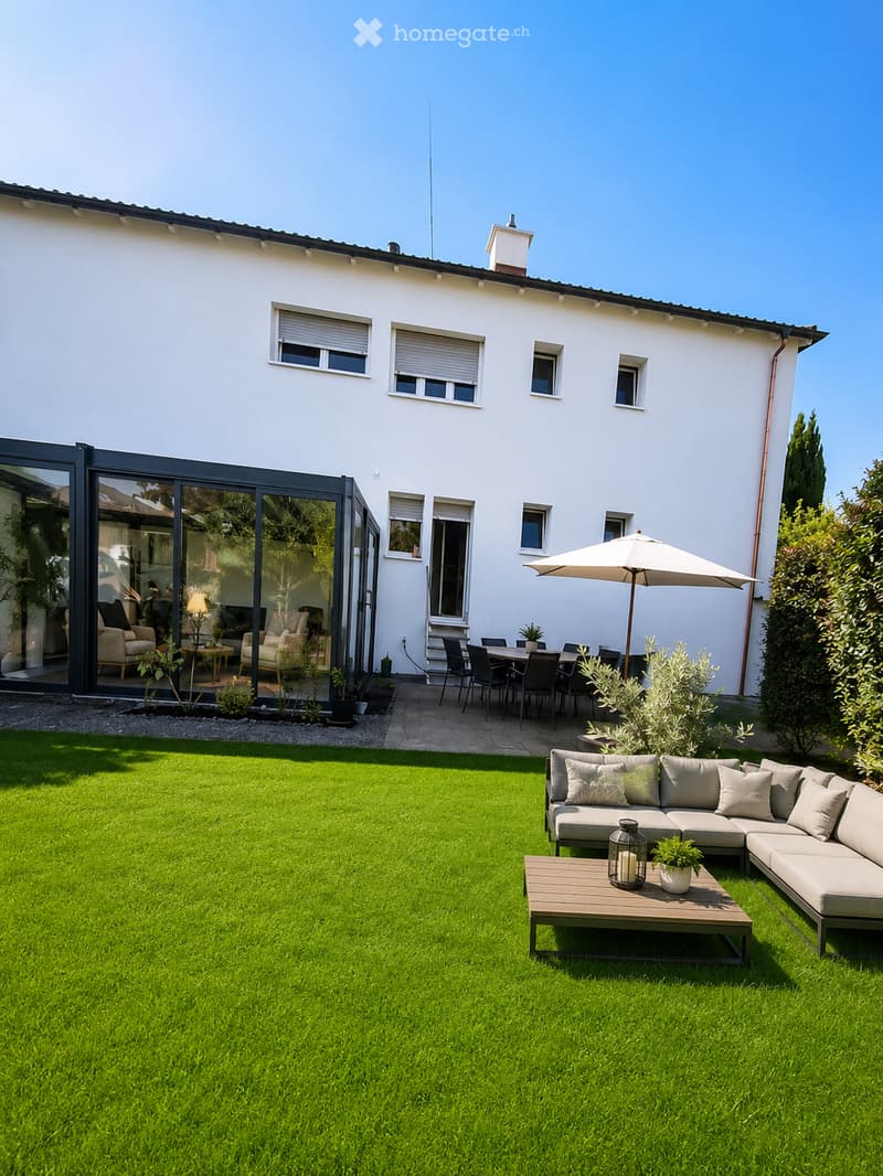 Well-maintained garden with a green lawn, a modern outdoor seating area featuring a sofa, coffee table, and umbrella, along with a dining set under a large umbrella near the house.