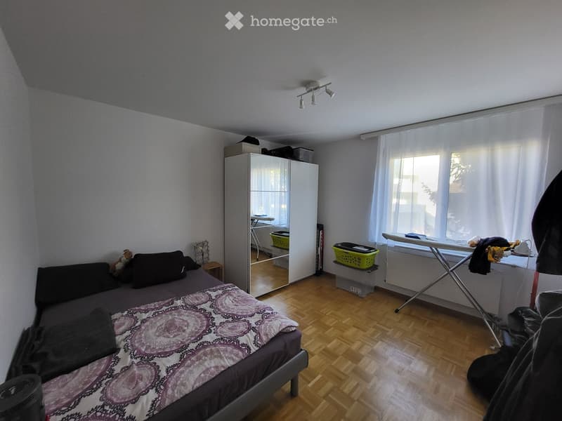 Bedroom with wooden flooring, a bed with dark bedding, a wardrobe, an ironing board, and a small stool.