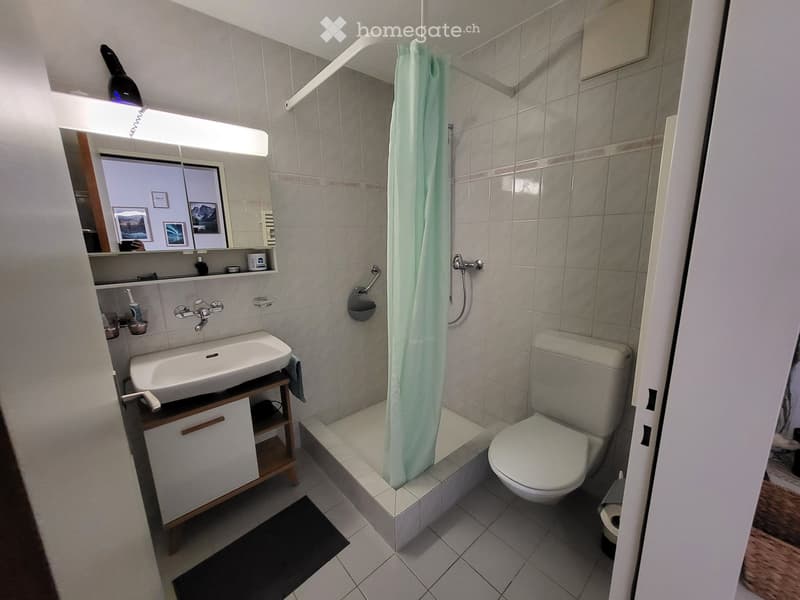 Modern bathroom with a white sink, toilet, mirror, and a shower area with a light green curtain. The room features tiled walls and floor, a small wooden cabinet under the sink, and a black bath mat.