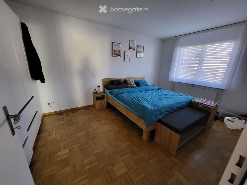 Cozy bedroom with wooden flooring, a bed with blue bedding, a nightstand, and a bench at the foot of the bed. The room features white walls, framed pictures, and a window with blinds.