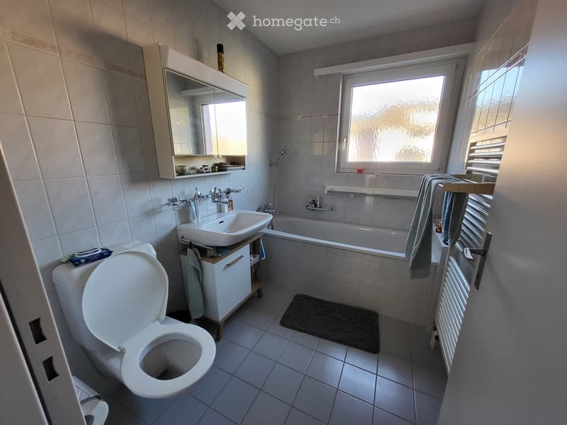 Modern bathroom with a toilet, sink, bathtub, mirror, radiator, and towel. The room features tiled walls and floor, a window with blinds, and a small cabinet under the sink.