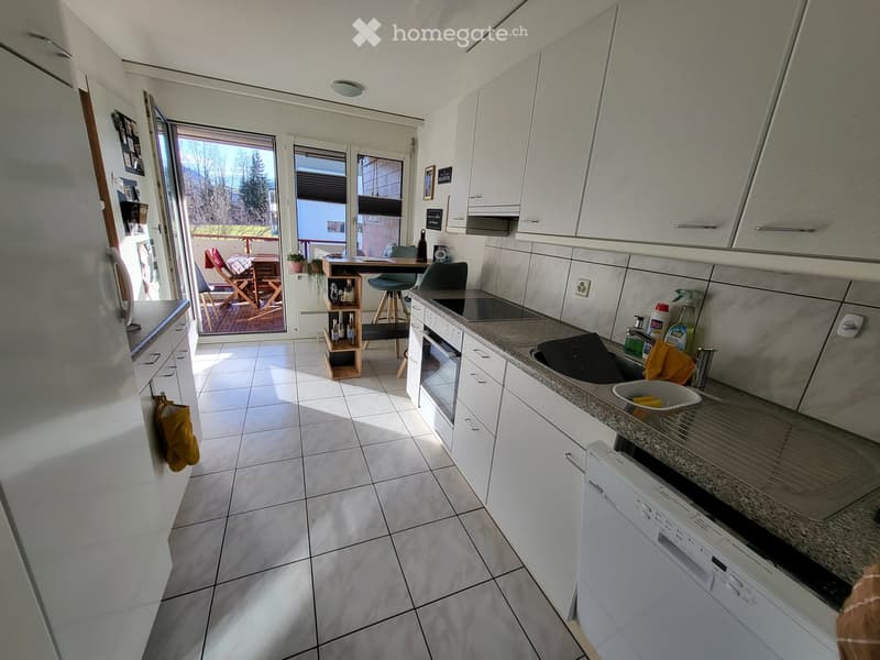 Bright, modern kitchen with white cabinets, granite countertops, and a large window offering a view of the outdoor area. Includes a refrigerator, oven, dishwasher, sink, and a small dining area with a table and chairs.