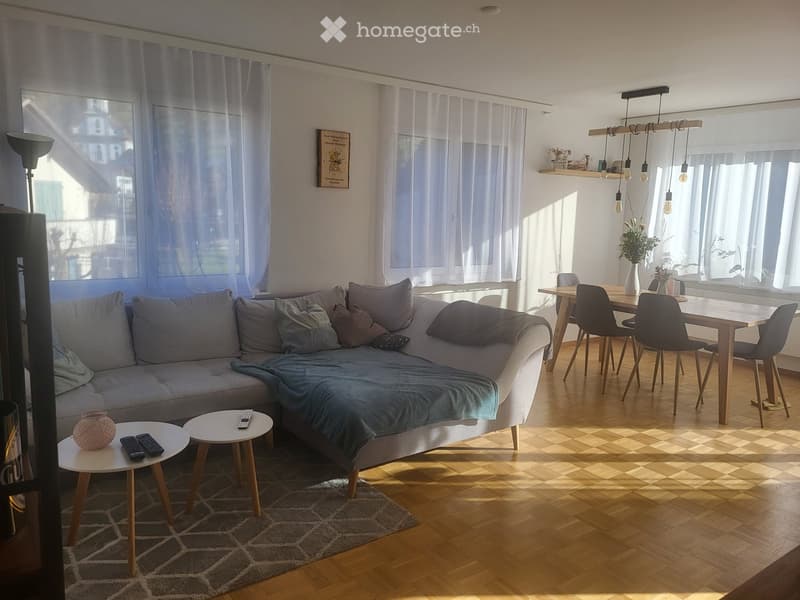 Bright living room with a grey sectional sofa, two small coffee tables, a dining table with four chairs, a floor lamp, and a vase on a shelf.