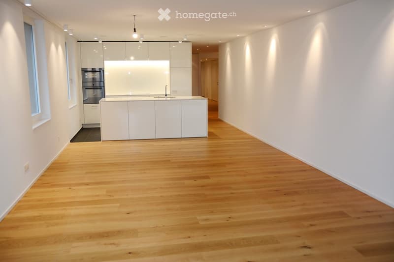 Empty room with light wooden flooring, white walls, and a small kitchen area with white cabinets and stainless steel appliances visible in the background.