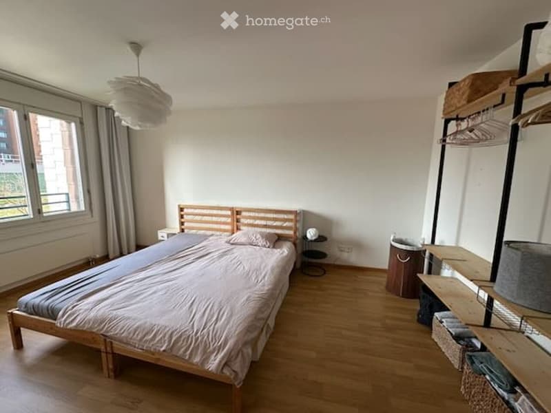 A modern, minimalist bedroom with a wooden bed, nightstand, small chair, and shelving unit. The room features light-colored walls, wooden flooring, and a large window with curtains.