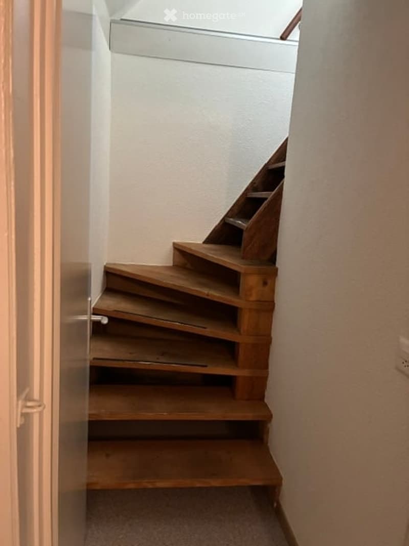 A narrow wooden staircase with a simple design, leading upwards. The walls are painted white, and the floor is carpeted.