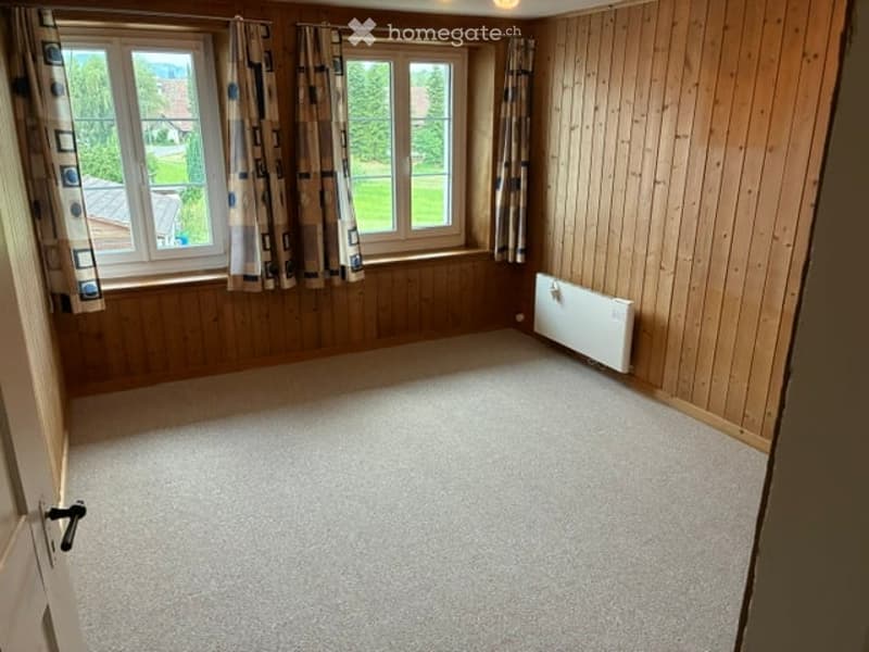 Empty room with wooden walls, carpeted floor, large windows with curtains, and a radiator on one wall.