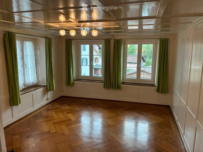 Empty room with wooden flooring, large windows with green curtains, and recessed lighting. No furniture or equipment present.
