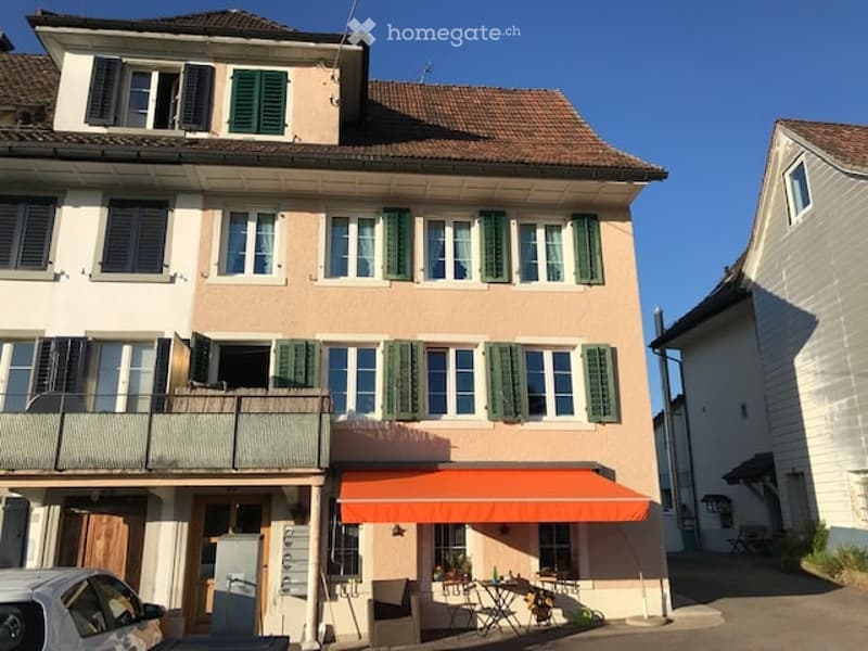 Two-story residential building with beige facade, green shutters, and a small balcony on the first floor. An orange awning covers the entrance area, and outdoor furniture is placed near the entrance.