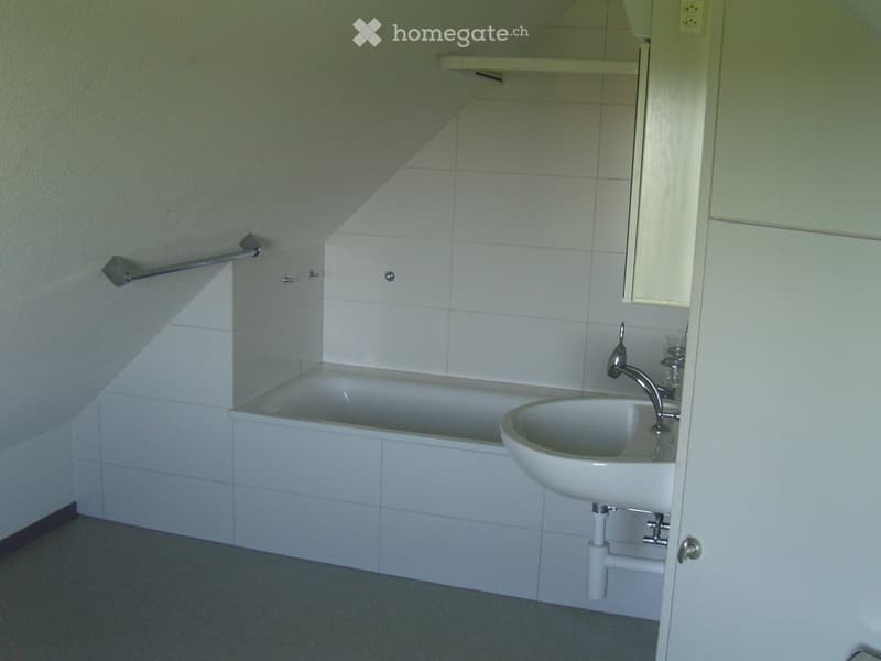 Bathroom with a bathtub, sink, faucet, mirror, and handrail. The walls are tiled, and the floor is grey.