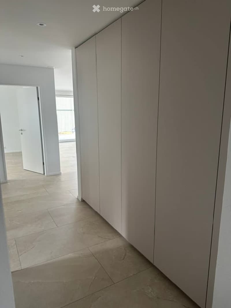 Empty hallway with beige tiled flooring, white walls, and built-in closet doors. Natural light streams in from an adjacent room.
