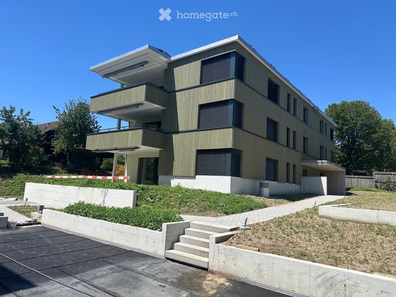Modern two-story house with green siding, large windows, and balconies. The house is surrounded by a landscaped area with concrete barriers and greenery.