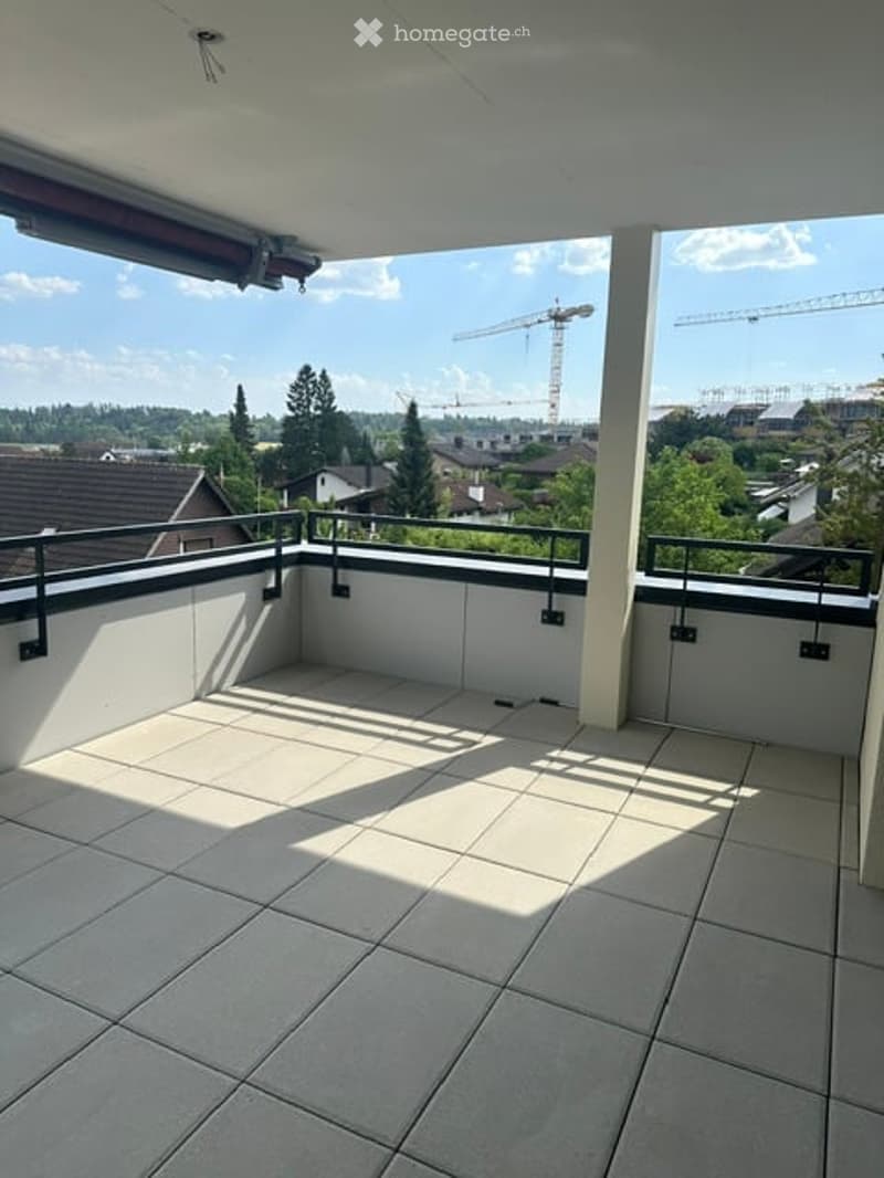Empty balcony with tiled floor, white walls, metal railing, and a view of a cityscape with construction cranes and trees.