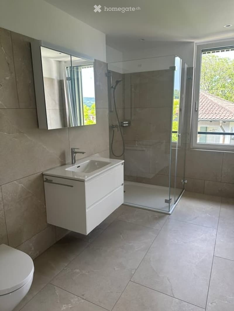 Modern bathroom with a white sink, large mirror, toilet, and a glass-enclosed shower. The space features beige tiles and a sleek, contemporary design.