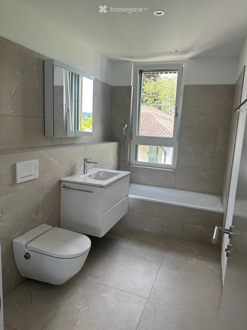 Modern bathroom with a white sink, toilet, bathtub, and a large window providing natural light. The walls are tiled in a neutral color, and there is a mirror above the sink.