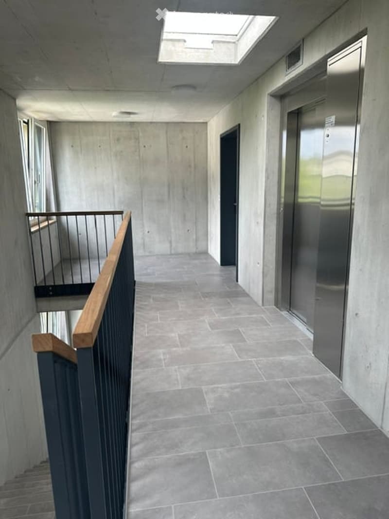 Modern hallway with concrete walls, tiled floor, elevator, and a small balcony area visible in the background.