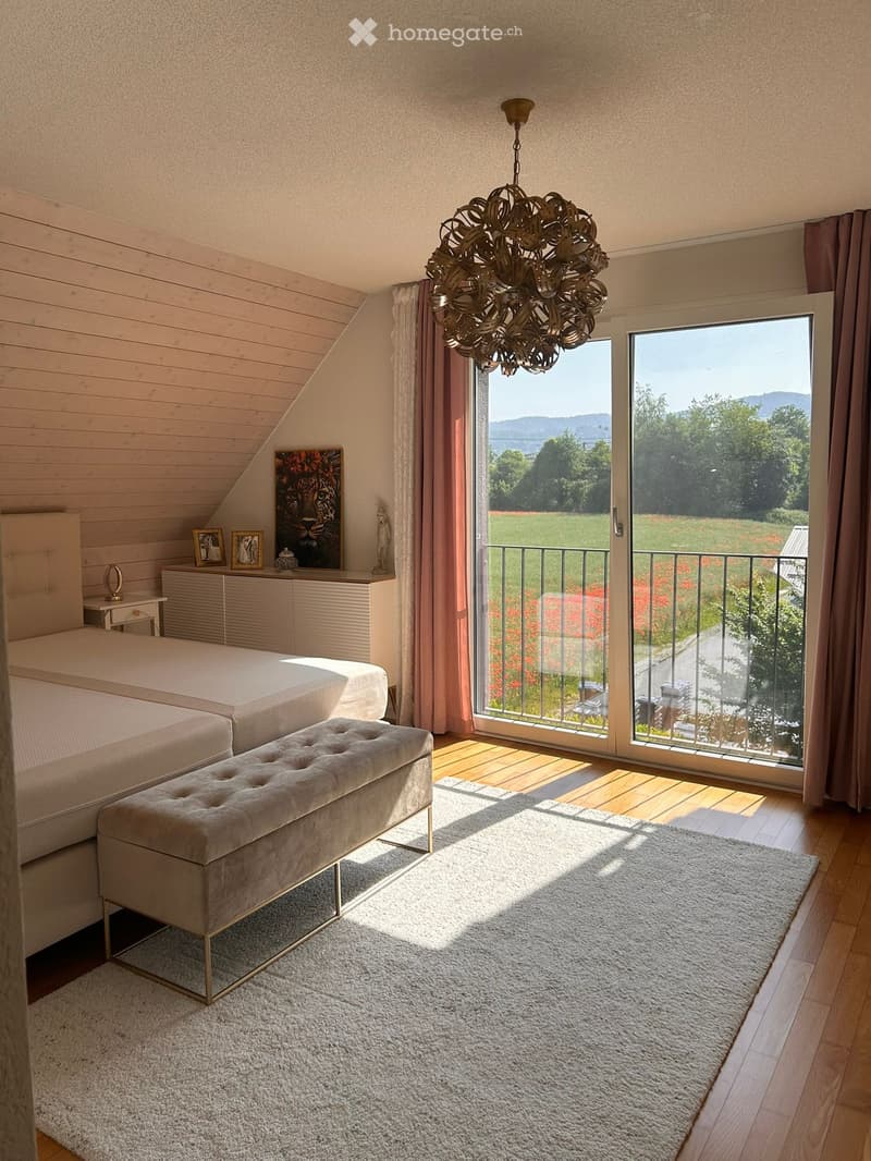 Bright bedroom with wooden slanted ceiling, large window with view, bed, bench, chandelier, picture frame, small table, and rug.