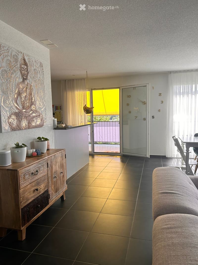 Modern living room with a sofa, coffee table, dining table, chairs, a wooden cabinet, plants, and a large Buddha wall art. The room features a sliding glass door leading to a balcony.