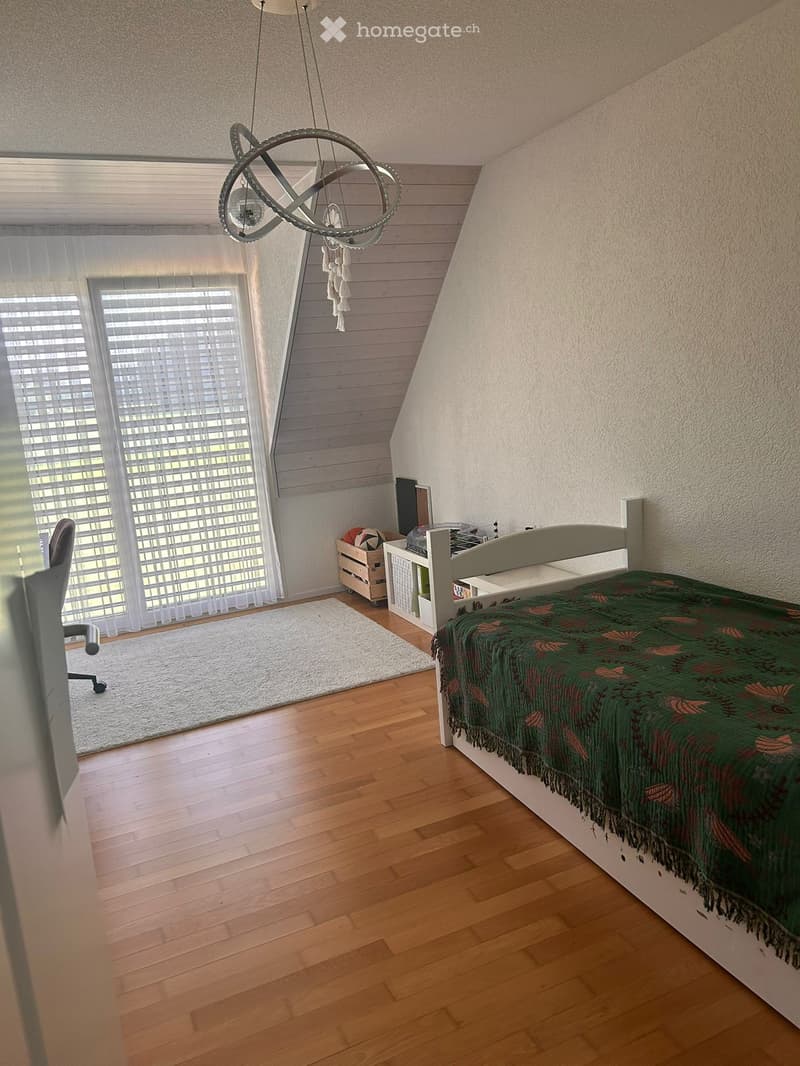 Cozy bedroom with wooden flooring, slanted ceiling, large window with blinds, bed with green patterned blanket, small table, chair, and decorative lamp.