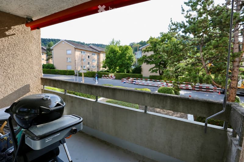 Balcony with a black grill, concrete railing, and a view of a street with buildings and trees.