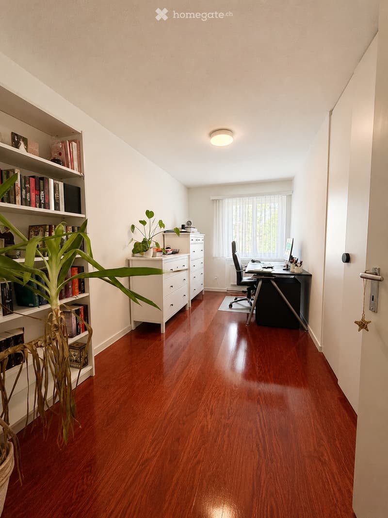 Well-lit study or home office with wooden flooring, white walls, a large window with blinds, a desk with a computer setup, a bookshelf filled with books, and a potted plant.