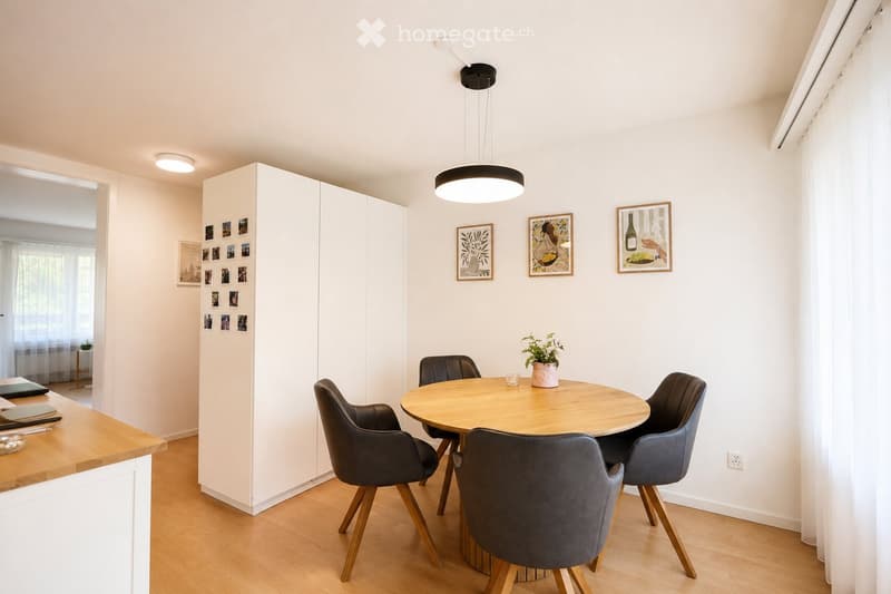 Modern dining area with a round wooden table surrounded by four black chairs, a pendant light fixture, white walls, and a built-in wardrobe with picture frames and a small plant on the table.