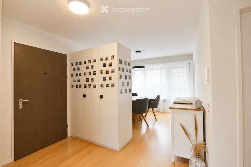 A modern hallway with a wooden door, a white cabinet with photo frames, a small dining area with chairs and a table, and a small white side table with a cup and a decorative vase.