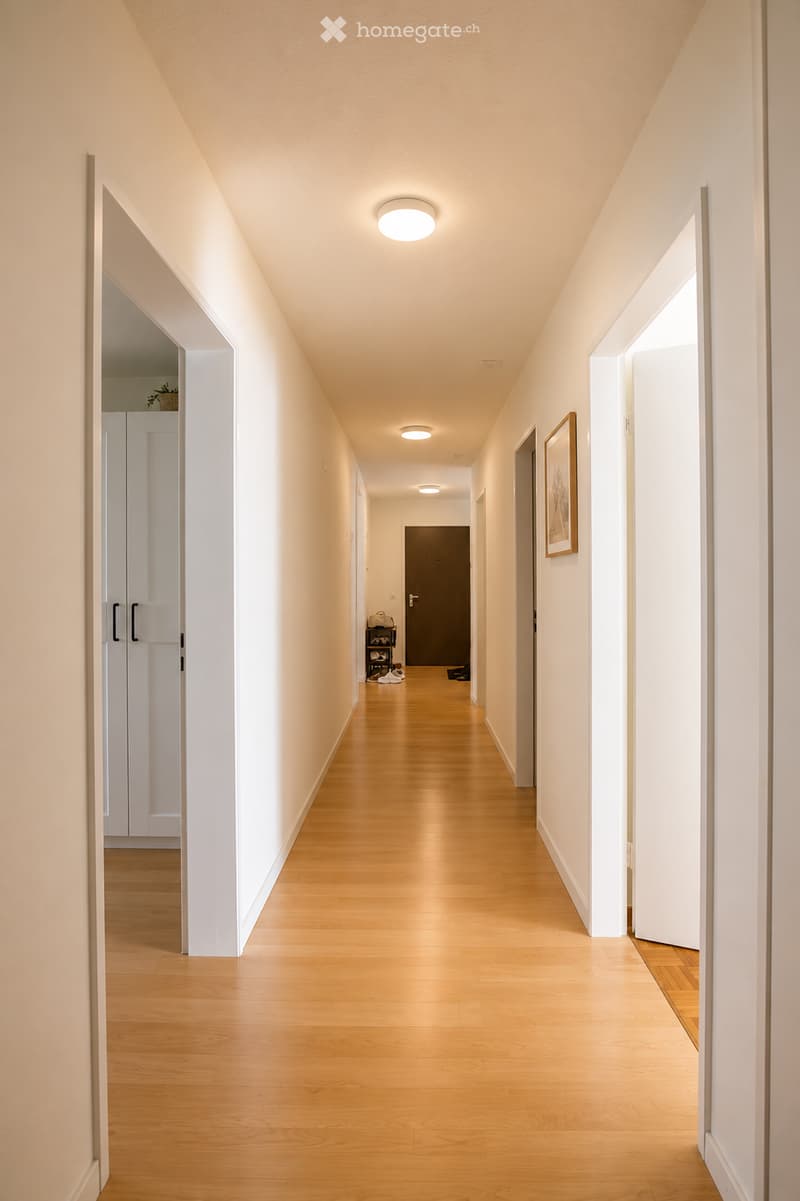 A clean, well-lit hallway with light wooden flooring, white walls, and recessed ceiling lights. Doors and a small shelf are visible along the corridor.