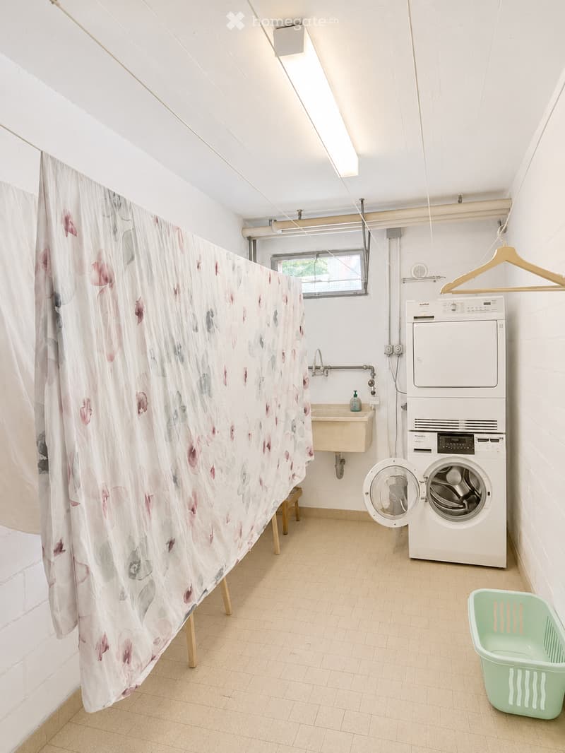 Bright bathroom with a washing machine, sink, and floral shower curtain. Includes a small stool and a laundry basket.