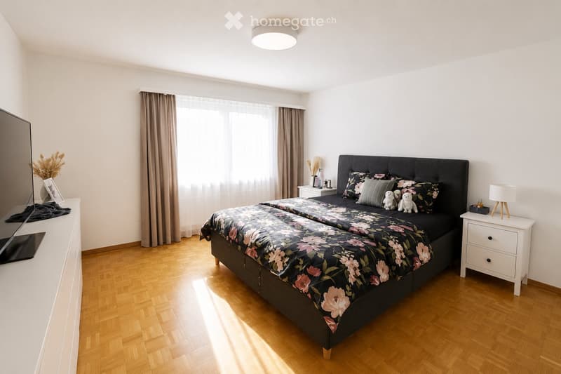 Modern bedroom with a large bed, nightstands, lamps, and a TV on a stand. The room features wooden flooring and large windows with curtains.
