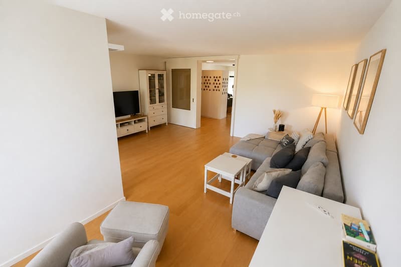 Modern living room with light wooden flooring, white walls, grey sofa, ottoman, coffee table, floor lamp, cabinet, and television.