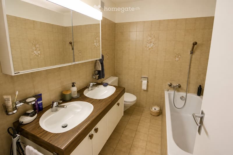 Modern bathroom with double sinks, a bathtub, a toilet, and a large mirror above the sinks. The countertop is made of wood, and various toiletries are neatly arranged.