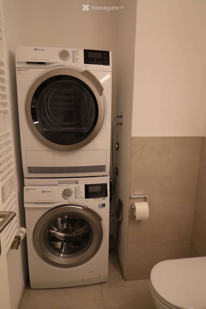 A compact bathroom with a stacked washing machine and dryer unit, a toilet, and a toilet paper holder.