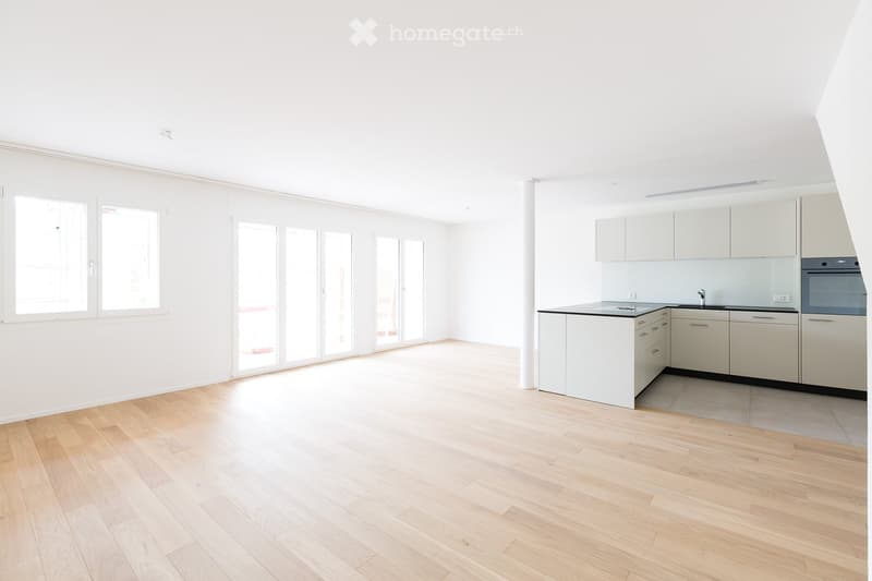 Bright, open-plan space with light wooden flooring, white walls, and large windows. A modern kitchen area features white cabinets, a sink, and an oven.