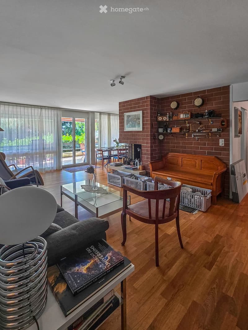Living room with wooden flooring, fireplace, sofa, coffee table, dining area, and large windows with curtains.