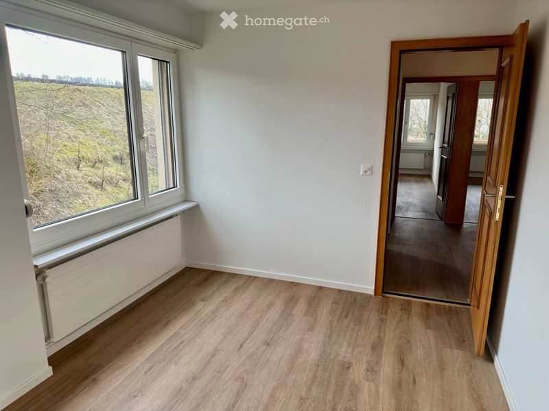 Empty room with wooden flooring, white walls, large window with view, radiator, and open doorway leading to another room.