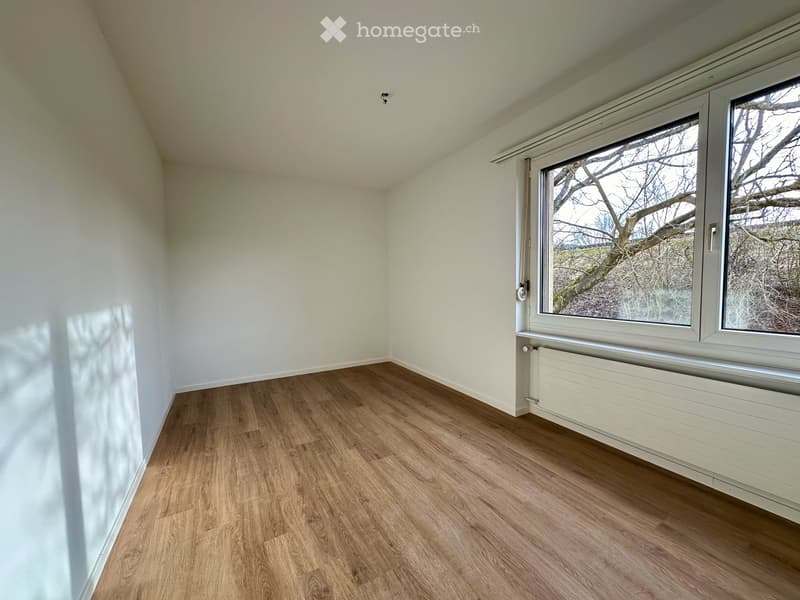 Empty room with wooden flooring, white walls, and large windows offering a view of trees outside.
