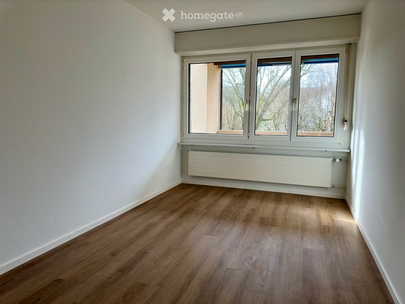 Empty room with wooden flooring, white walls, large windows, and a radiator below the windows.