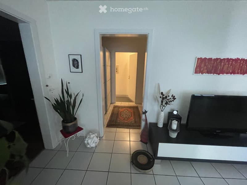 A modern living room with tiled flooring, a television on a stand, a potted plant on a small table, and a doorway leading to another room.