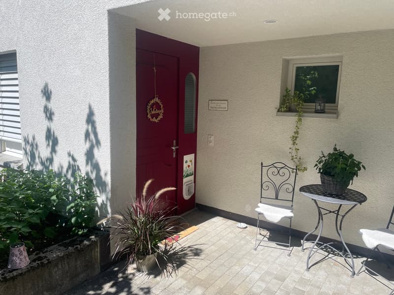 A small outdoor seating area with a metal chair, round table, and potted plants, adjacent to a red front door and window with a small plant on the sill.