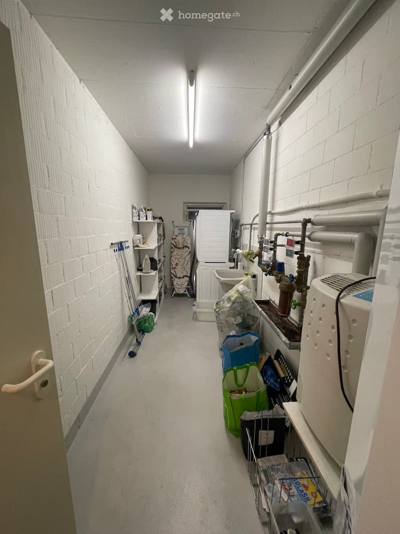 A narrow, white-walled corridor with exposed pipes and a small utility area at the end, containing a sink, shelves, and cleaning supplies.