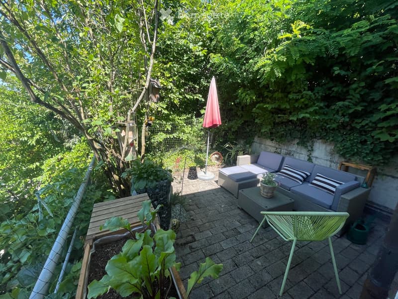 A lush, green outdoor patio area with a grey wicker sofa, a small table, a green chair, and a closed umbrella. The space is surrounded by trees and plants, creating a serene and private atmosphere.