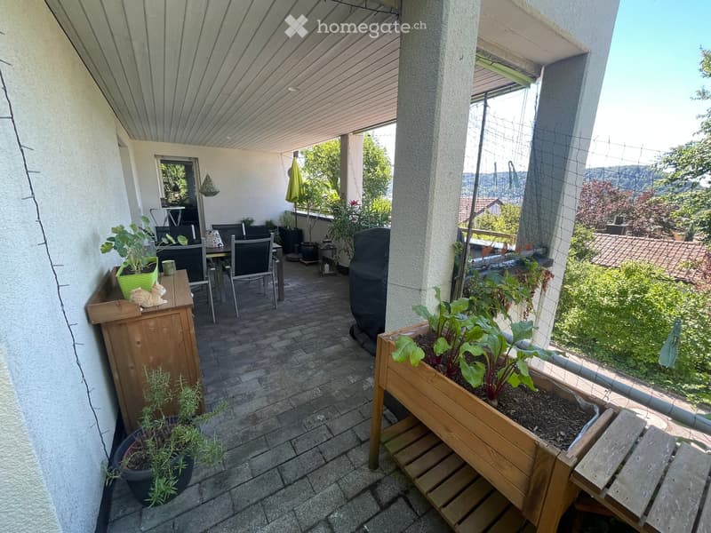 Balcony with a covered area, tiled floor, outdoor furniture including a table and chairs, potted plants, and a wooden planter box with vegetables.