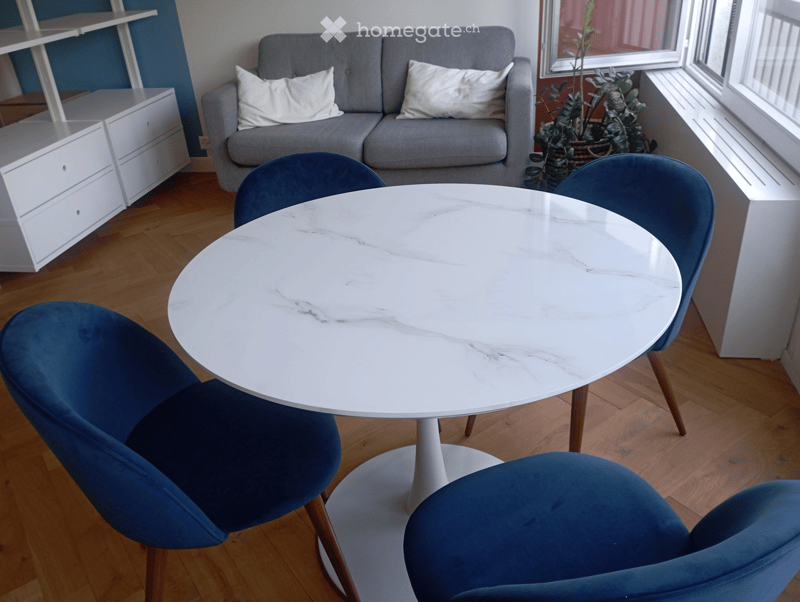 Modern living room with a grey sofa, white marble table, four blue chairs, white cabinet, and a potted plant near the window.
