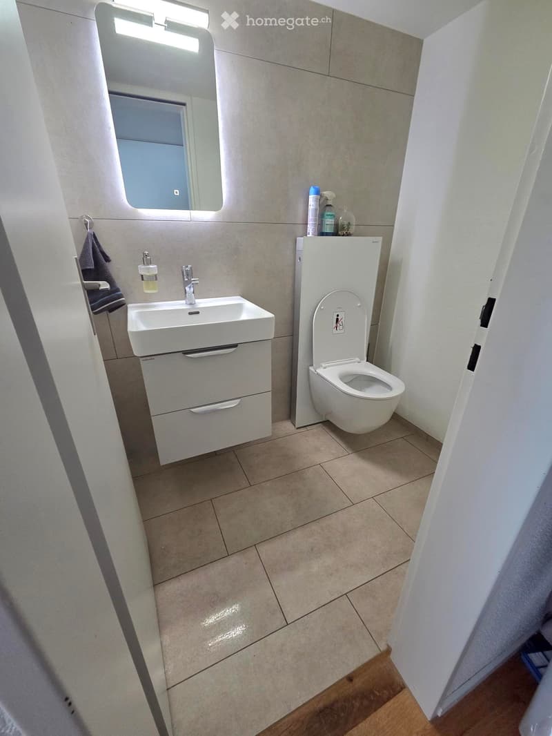 Modern bathroom with a white sink, toilet, and mirror. Beige tiled floor and walls, with a towel and bottle on the sink counter.