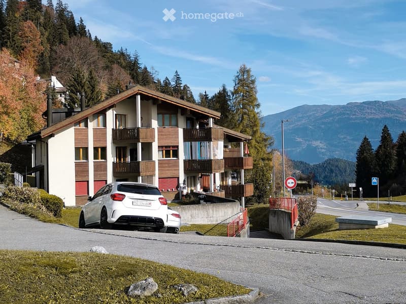 A multi-story residential building with balconies, surrounded by trees and mountains, with a parked car in front.