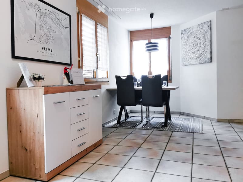 Bright room with tiled floor, white walls, and large windows. Features a wooden cabinet with drawers, a dining table with four chairs, and decorative items on the cabinet.