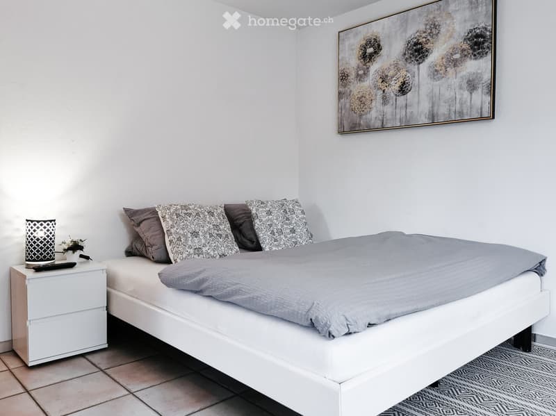 Modern bedroom with a white bed, grey bedding, decorative pillows, a nightstand with a lamp, a remote control, a small flower vase, and a large abstract painting on the wall.