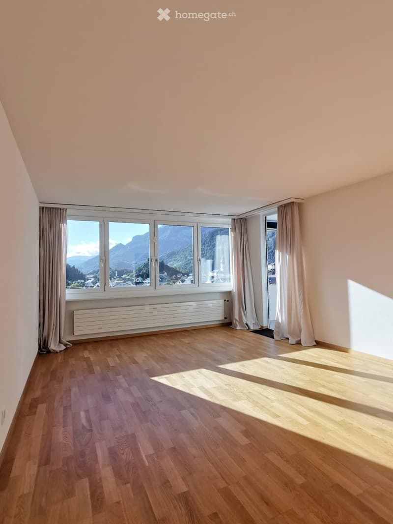 Empty room with wooden flooring, large windows with curtains, and a radiator below the windows.